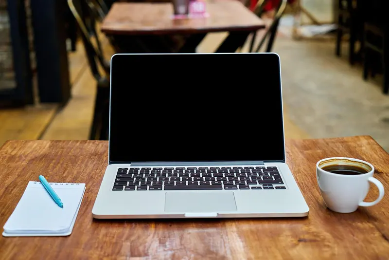 Aufgeklappter Laptop mit dunklem Bildschirm auf Holztisch, daneben Kaffeetasse und Notizblock mit Kugelschreiber
